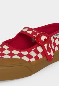 Chaussure à enfiler à motif damier rouge et blanc avec une bride à boucle rouge et une semelle en caoutchouc marron, logo Vans sur le côté.