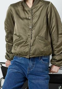 Olivgrön bomberjacka med satinfinish, tryckknappar, ribbad krage och sidofickor, i kombination med blå denimjeans.