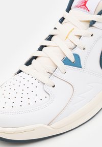 Baskets blanches avec boîtier perforé, lacets beiges, accents bleus et cuir texturé. Présente un logo sur la languette et des détails cousus.