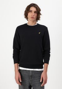 Zwarte sweater met ronde hals en geribbelde manchetten, voorzien van een klein gouden logo op de borst. Gedragen over een witte gelaagde shirt.