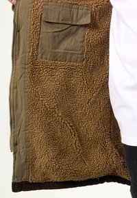 Giacca marrone con interno in pile, esterno verde oliva e tasca sul petto. Presenta un interno morbido e testurizzato e un orlo a coste.