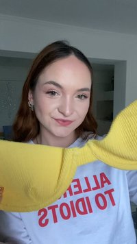 Ung kvinna med ljus hy och brunt hår håller en ljusgul bh, klädd i en vit tröja med röd text inomhus.