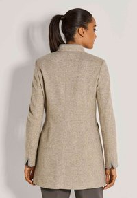 Vrouw met donker haar in een staart, gekleed in een beige lange blazer, gezien vanuit de rug, staand tegen een lichte muur.