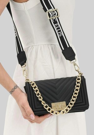 Donna in abito bianco che tiene una borsa nera trapuntata con catena dorata e tracolla regolabile nera e bianca con marchio.