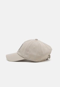 Cappello beige in cotone con visiera curvata, cinturino regolabile e un piccolo logo ricamato sul davanti. Superficie testurizzata, design a sei pannelli.