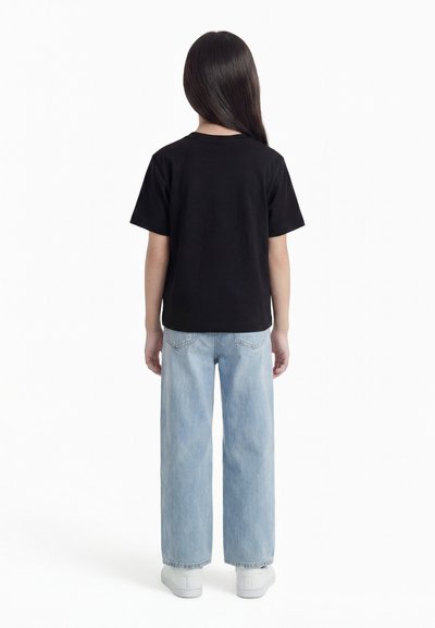 Παιδί με μακριά μαλλιά που φοράει μαύρο κοντομάνικο πουκάμισο, ανοιχτό μπλε τζιν παντελόνι με φαρδιά μπατζάκια και λευκά αθλητικά παπούτσια, στέκεται γυρισμένο από την πλάτη.