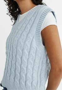 Hellblauer, gestrickter ärmelloser Pullover mit V-Ausschnitt, mit Zopfmuster und gerippten Kanten, über einem weißen Hemd getragen.