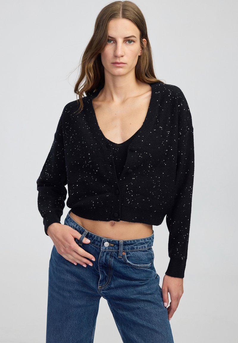 Donna con lunghi capelli castani, che indossa un cardigan corto nero con bottoni e macchie bianche, abbinato a jeans a vita alta blu, in piedi davanti a uno sfondo neutro.