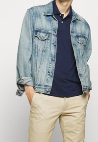 Chaqueta de mezclilla azul claro con dos bolsillos en el pecho, cierres de botones plateados y mangas enrolladas, llevada sobre una polo oscura y pantalones beige.