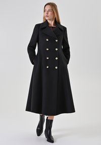Schwarzer, doppelt geknöpfter Mantel mit goldenen Knöpfen, großen Revers und Seitentaschen. Ausgestelltes A-Linien-Design, das bis zur Mitte der Wade reicht. Strukturierter Stoff.