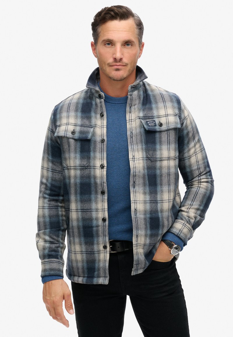 Camicia di flanella a quadri blu e crema, con chiusure a bottoni, due tasche sul petto e un colletto rialzato. Indossata sopra un maglione blu a texture.