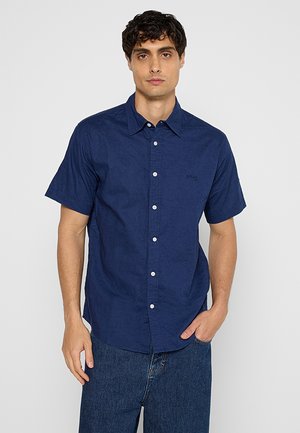 Korte mouwen, donkerblauwe linnen shirt met een knoopsluiting, puntige kraag en een klein logo op de borst, gecombineerd met donkere spijkerbroeken.