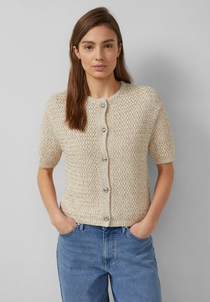 Vrouw met steil bruin haar, een beige korte mouw gebreid vest met knopen dragend, handen in de zakken, tegen een effen grijze achtergrond.