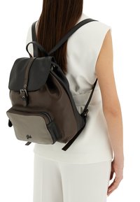 Zaino in pelle nera e marrone con una finitura testurizzata, chiusura a pattina, tasca frontale con zip e spallacci regolabili. Logo circolare alla base.