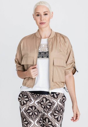 Frau mit kurzen blonden Haaren, die eine beigen Kurzarm-Jacke mit Reißverschluss über einem weißen, gemusterten Oberteil und gemusterten lockeren Hosen trägt.