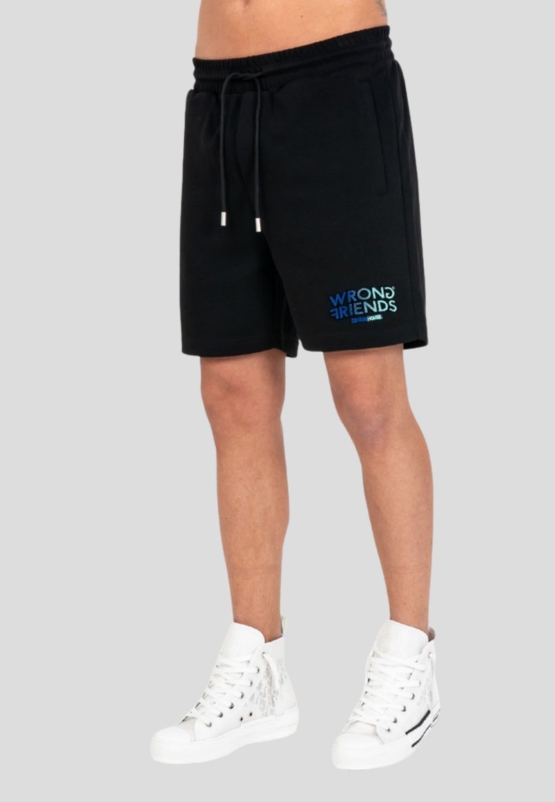 Black drawstring shorts with "WRONG FRIENDS" text logo on left leg, worn with white high-top sneakers on a plain background.