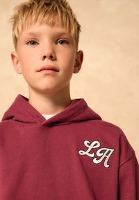 Felpa con cappuccio color bordeaux con una tasca frontale, caratterizzata da un ricamo bianco "LA" sul petto sinistro, realizzata in morbido cotone con una finitura testurizzata.