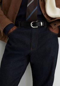 Mand iført mørkeblå jeans med orange syninger, sort bælte med sølvspænde, brun jakke, blå skjorte og stribet brun og blå slips.
