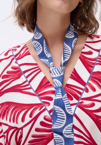 Rood-wit gestreepte blouse met een blauwe stropdas met witte bladerenmotieven. De stof heeft een gladde textuur en een V-hals ontwerp.
