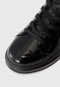 Tenis din piele lacuită neagră, cu vârful rotund, șireturi negre texturate și o suprafață netedă, reflectorizantă. Include o talpă din cauciuc negru și gromete.