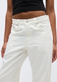 Κοντινό πλάνο ενός ατόμου που φοράει ψηλόμεσα λευκά τζιν με ασημένιο κουμπί και καρφιά, σε συνδυασμό με ένα μαύρο κοντό τοπ.