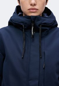 Jeune personne portant une veste à capuche bleu marine avec des cordons noirs et un col partiellement zippé avec une tirette visible.