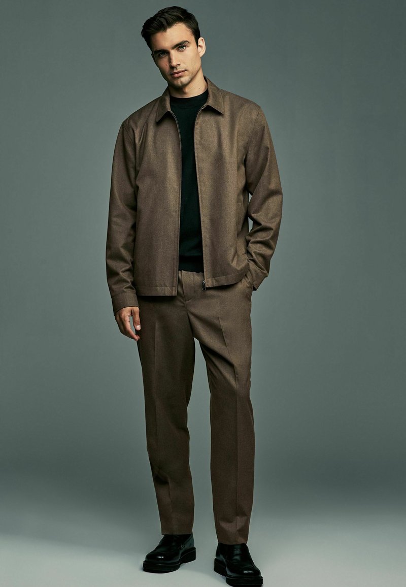 Braune Jacke mit Kragen über einem schwarzen Hemd, kombiniert mit braunen Hosen und schwarzen Schuhen. Glatte Textur, taillierte Passform und minimalistisches Design.