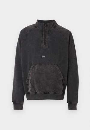 Άνδρας με μαύρο καπέλο, μαύρο φούτερ με φερμουάρ στο λαιμό, γκρι παντελόνι cargo, μαύρα παπούτσια, κρατώντας μεγάλη γκρι επενδεδυμένη τσάντα στο αριστερό χέρι.