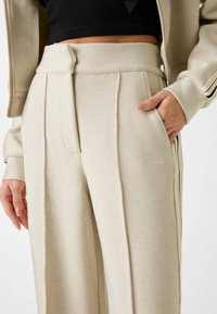Beige ribgebreide broek met een hoge taille, verticale plooien en zijzakken. Bevat subtiele geborduurde "Guess" logo's en zwart gestreepte accenten.