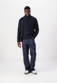 Marineblå høyhalset genser, strukturert strikk; mørke kamuflasje cargo-bukser med sidelommer; hvite og grå joggesko; svart skulderveske.