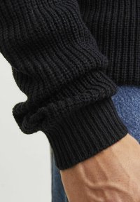 Black knit sweater cuff with ribbed texture, featuring a rolled edge. Close-up shows the soft fabric and hand detail.