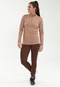 Helle braune Langarmbluse mit einem Reißverschluss-Kragen, kombiniert mit dunkelbraunen Leggings mit Seitentaschen. Schwarze Sportschuhe runden das Outfit ab.