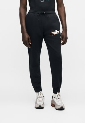 Osoba ubrana w czarne spodnie dresowe Nike z pomarańczowym logo i białe buty sportowe Nike stojąca na jednolitym białym tle.