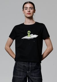 Schwarzes Baumwoll-T-Shirt mit einem grünen Alien-Graphic auf einem Papierflugzeug vor einem sternenverhangenen Hintergrund; kurze Ärmel und lockere Passform.