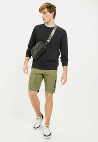 Schwarzer Sweatshirt, grüne Cargo-Shorts und weiße Turnschuhe mit marineblauen Akzenten. Eine kleine Umhängetasche wird über der linken Schulter getragen.