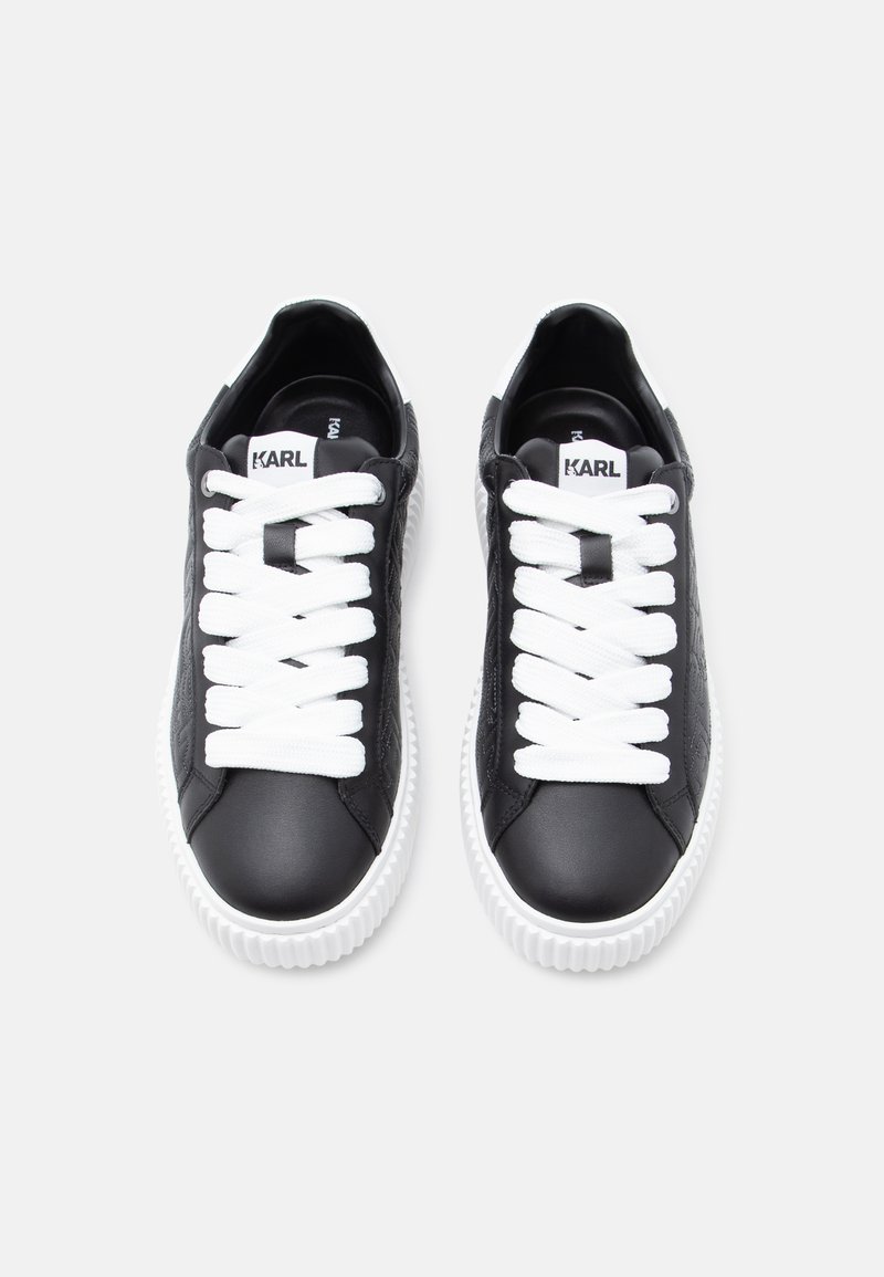 Zapatillas negras de cuero con cordones blancos, punta redonda, laterales texturizados y suelas de goma blancas. Etiqueta con logo en la lengüeta. Diseño plano y casual.