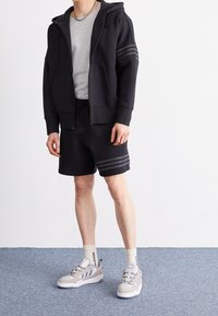 Svart zip-up hoodie med grå ränder på ärmarna, grå t-shirt, svarta shorts med ränder, och grå sneakers med blå accenter.