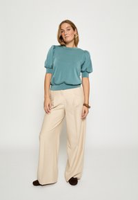 Morski zielony top z bufkami w połączeniu z beżowymi, szerokimi spodniami. Gładka tkanina o luźnym kroju. Brązowy dodatek na nadgarstku i ciemne buty.