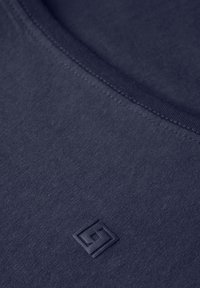 Marinblå tygtröja med rundad halsringning och en texturerad logotyp i nedre hörnet. Materialet verkar mjukt med en slät yta.