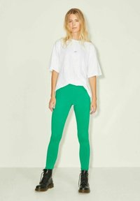 Zöld leggings, amely szoros szabású, rugalmas anyagból készült, laza fehér pólóval és fekete fűzős csizmával párosítva.