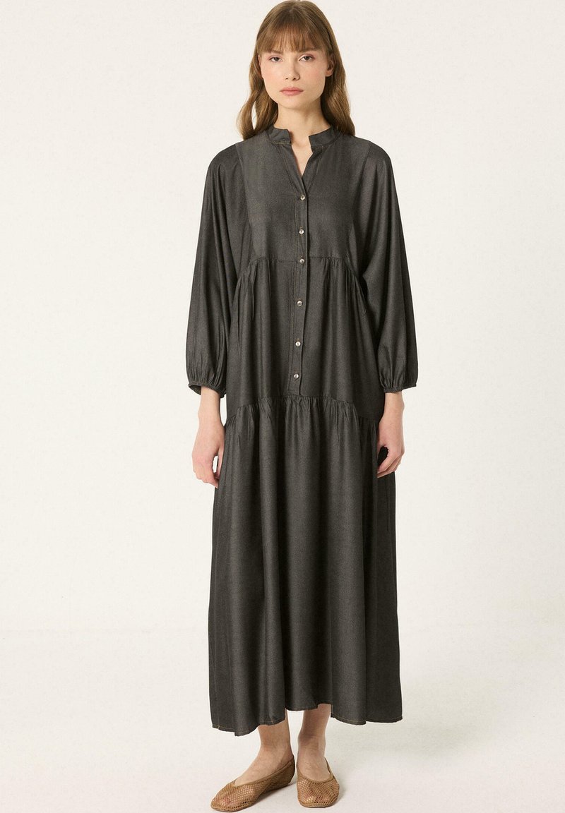 Femme portant une robe maxi noire à manches longues, boutonnée avec une taille froncée et des chaussures plates marron, debout devant un fond uni.