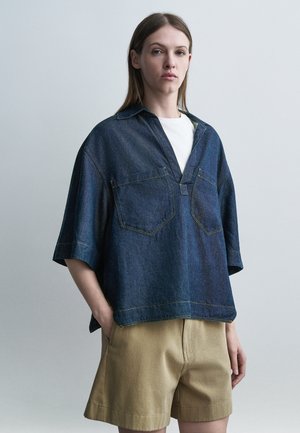 Nő, aki túlságosan nagy kék farmeringet visel fehér felső és bézs rövidnadrág fölött, egy kézzel a zsebében áll egy egyszínű háttér előtt.