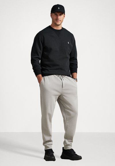 Μαύρη μπλούζα με στρογγυλή λαιμόκοψη, γκρι φόρμες και μαύρα αθλητικά παπούτσια. Η μπλούζα έχει ένα μικρό λογότυπο, ενώ οι φόρμες διαθέτουν ελαστική μέση και μανσέτες.