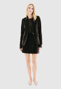 Femme portant un blazer noir, une jupe noire et des ballerines pointues au motif serpent, se tenant devant un fond blanc uni.