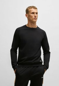 Sweat-shirt noir à manches longues et col rond, avec une texture lisse et des manches raglan. Présente un petit logo sur l'ourlet inférieur.