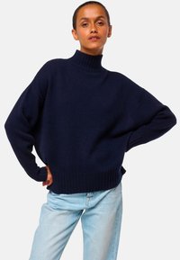 Femme portant un pull col montant bleu marine et un jean bleu clair, posant les mains sur les hanches devant un fond blanc uni.