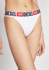 Bela tangica z črno-rdečim pasom z oznako "DIESEL". Mehak material, minimalna pokritost in gladka tekstura.