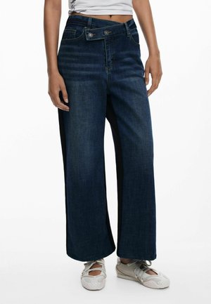 Wijd gesneden jeans van donkerblauw denim met een lichte vervaging, met een knoopsluiting in de taille en zwarte zijpaneelaccenten. Gedragen met lichte sneakers.