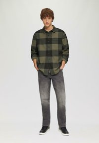 Groen en zwart geblokt flanellen shirt met een geknoopte voorkant en een borstzak, gecombineerd met vervaagde grijze spijkerbroeken en zwarte sneakers.