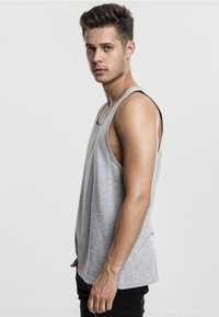 Grå ermeløs tank topp laget av bomull, med en avslappet passform og myk tekstur, kombinert med svarte bukser. Modellen poserer i profil.